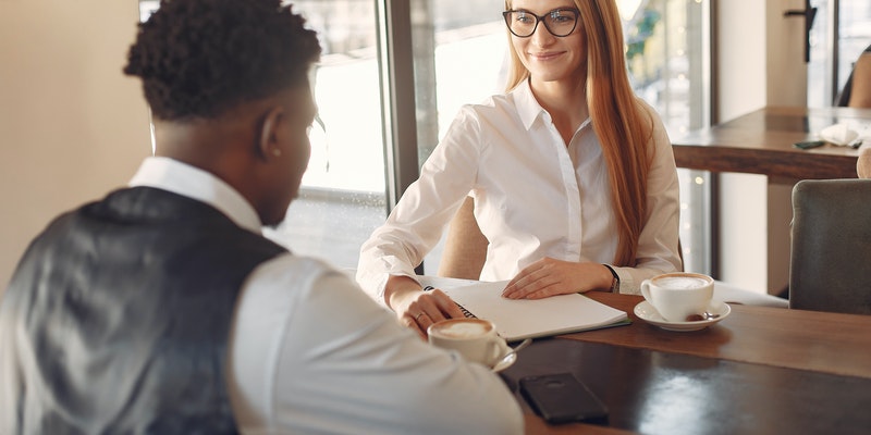 A man and woman seated at a desk across from each other. Both are dressed in professional attire and appear to be engaging in a job interview.